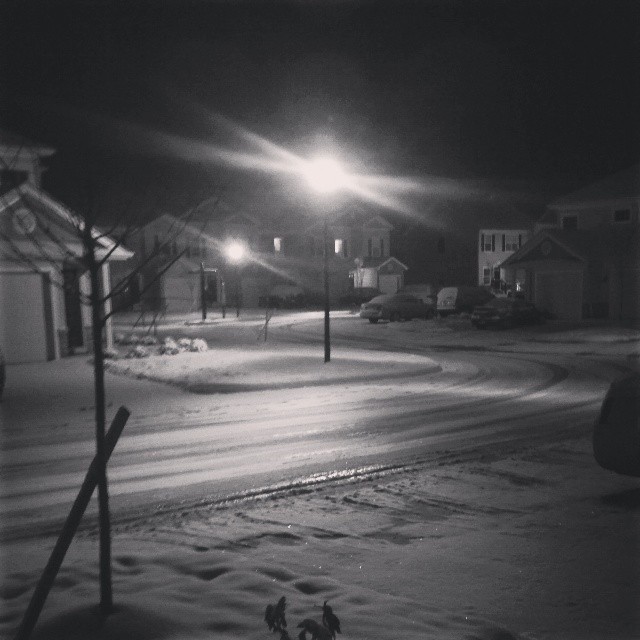 Black and white, snow covered suburban at night with centered light post flaring